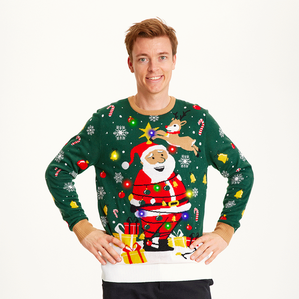 Een man met zijn handen langs zijn zij die een groene kersttrui met kerstman en lichtjes draagt.